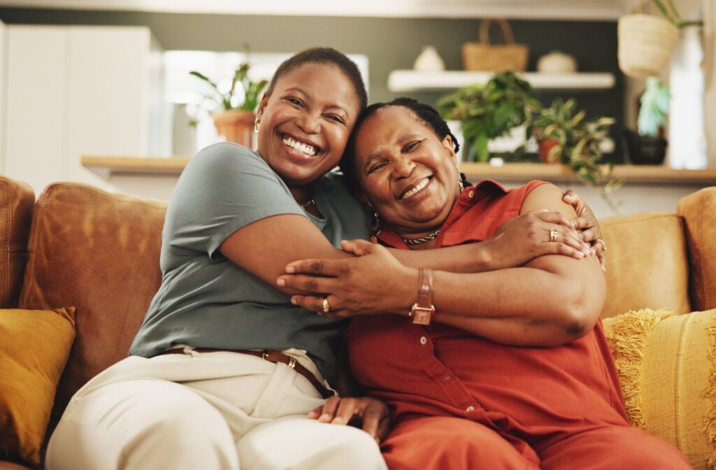 An adult and their older parent happily hug each other on a couch during a visit to their parent’s assisted living home