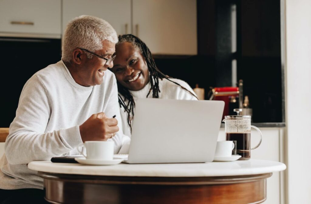 An older senior couple laugh together over coffee while researching senior living options for their retirement on a laptop
