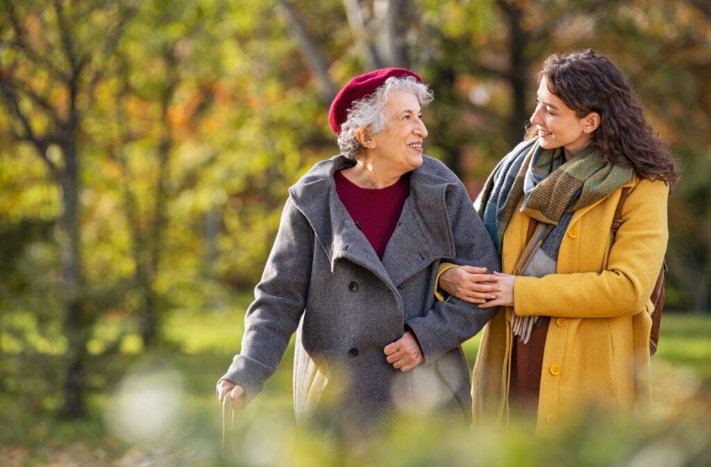 An adult, wearing a yellow coat, walks with their senior parent, arm-in-arm, in a park during autumn and discusses assisted living options.
