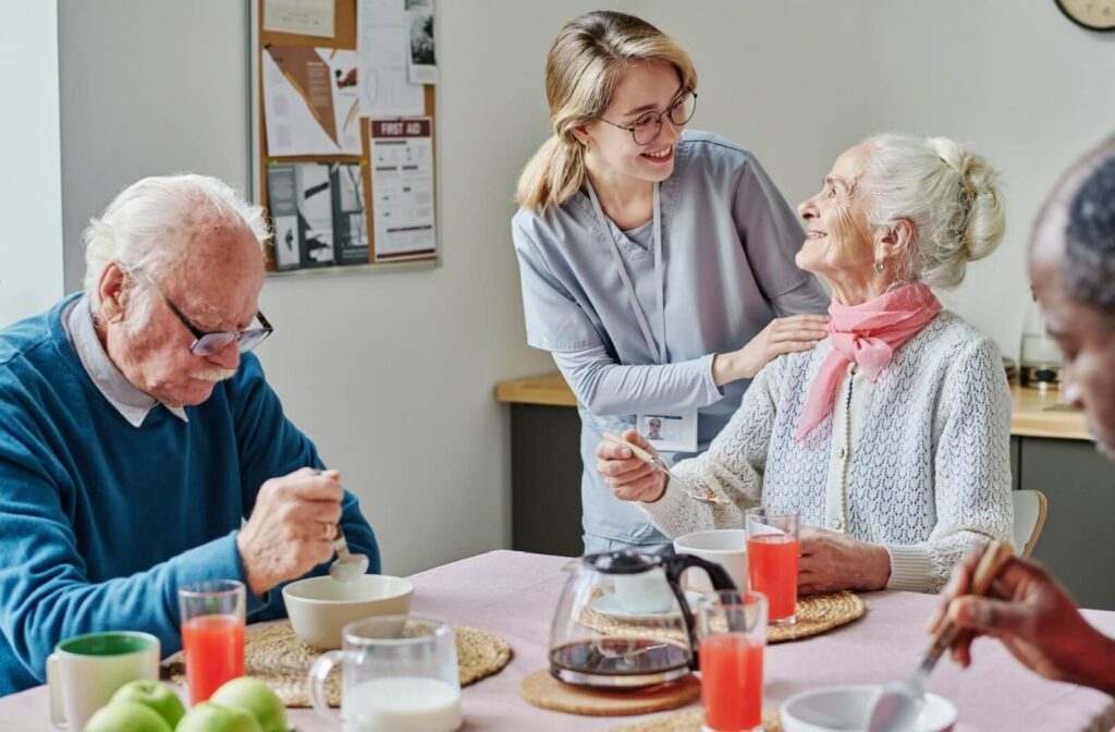 An older adult looks up and smiles during breakfast as a happy caregiver touches their shoulder and checks in