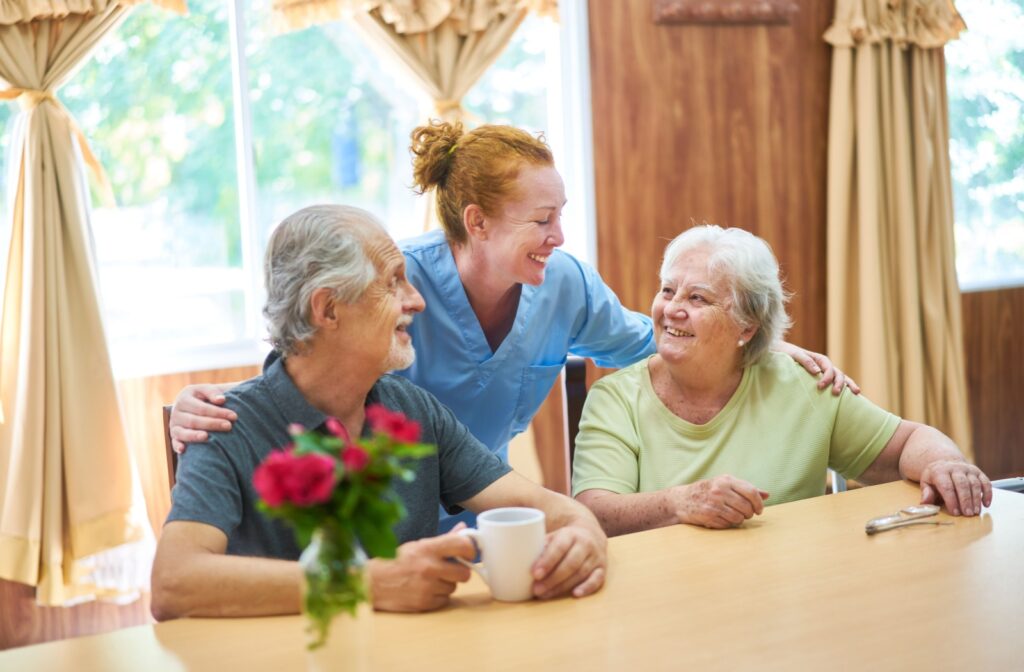 A smiling caregiver checks in on two happy residents in assisted living during a coffee hour in a communal dining area
