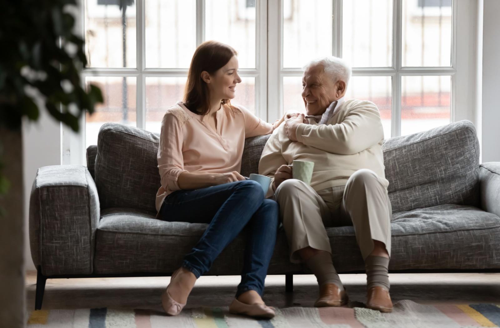 A younger adult sits on the couch and has a conversation with their senior loved one over a cup of coffee.
