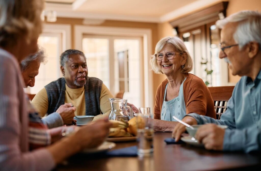 Five older adults smile while sitting around a breakfast table in a warm, sunlit dining room in assisted living
